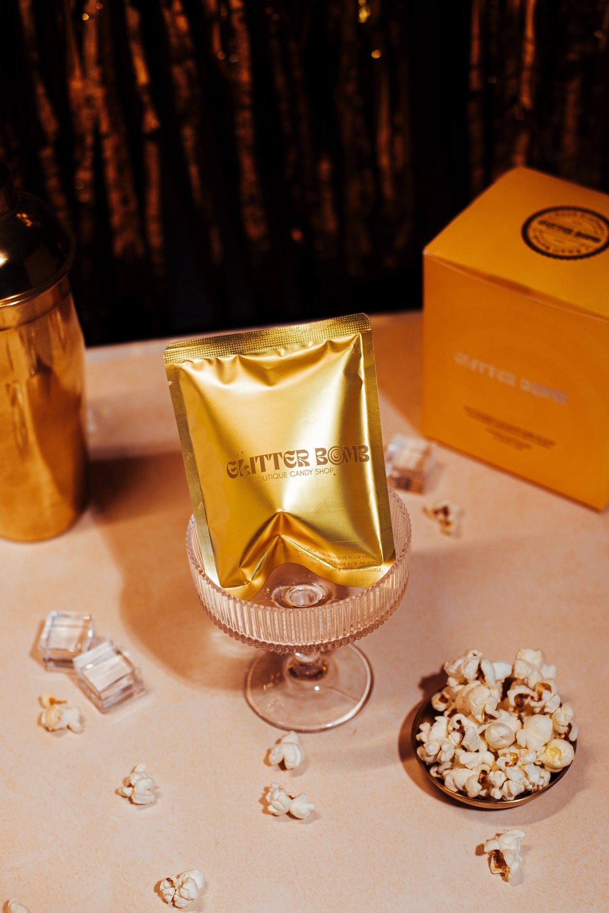 A gold-colored packet labeled 'Gitter Bomb' is displayed on a glass surface with scattered glitter and a box in the background.